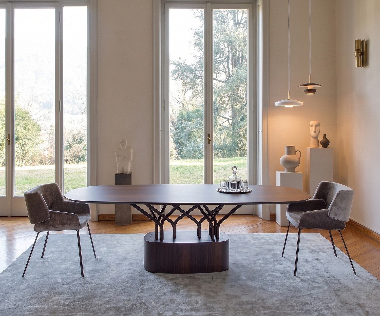 Ovaler al2 wood-oo Esstisch aus Holz im modernen Esszimmer mit zwei grauen Samtsesseln vor großen Fenstern.