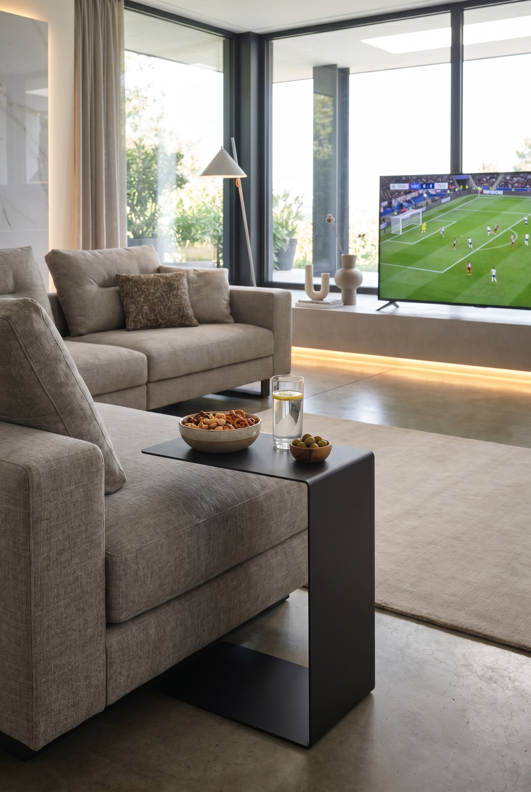 Ein moderner Marelli Mini Sofa Beistelltisch in C-Form, dessen Ablagefläche mit einem Getränk und Snacks über die Armlehne eines gemütlichen Sofas reicht.