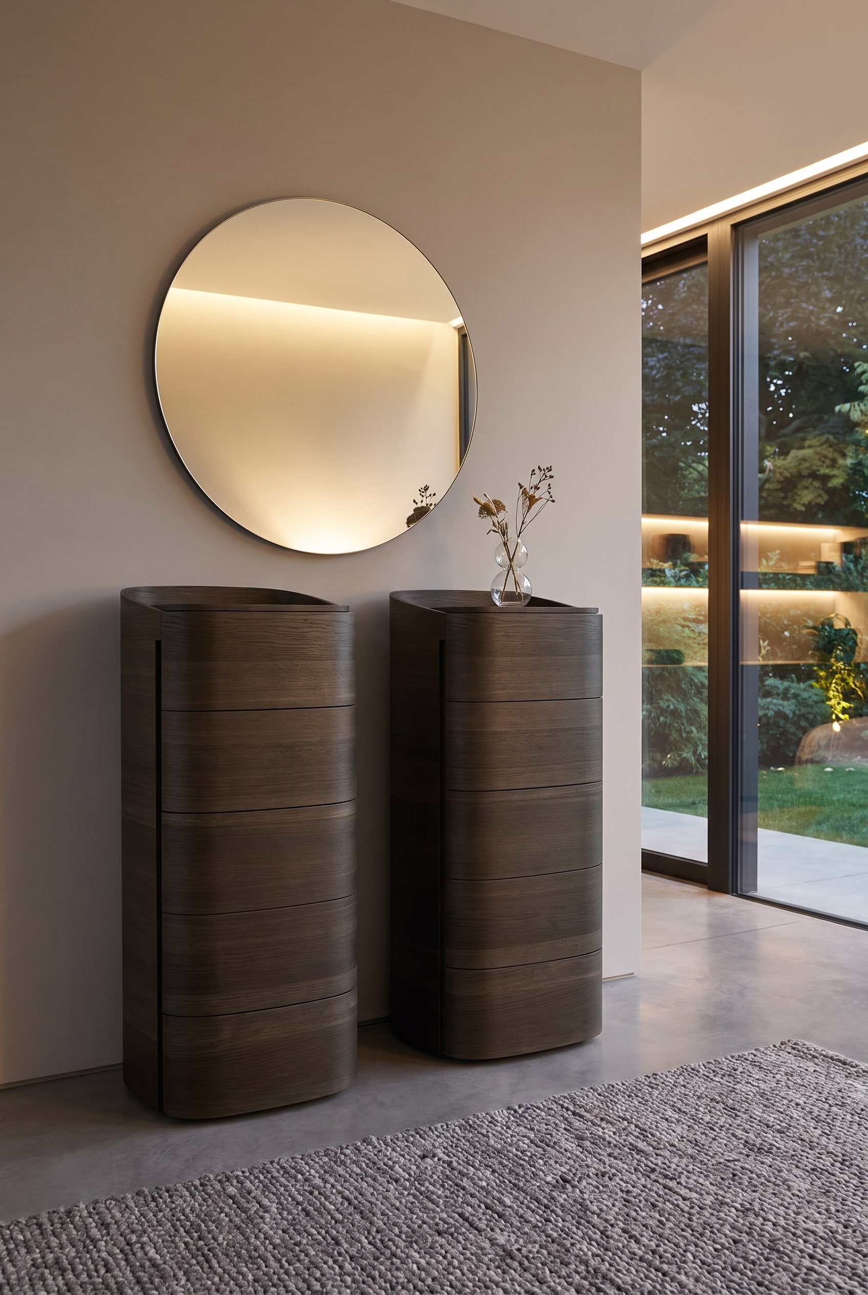 Hohe, abgerundete Kommode aus dunklem Holz, darüber ein großer, minimalistischer Rundspiegel an der Wand, davor eine kleine Vase