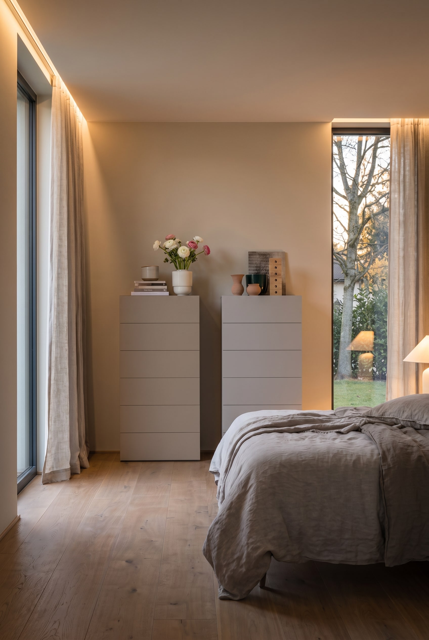 Zwei greige Hochkommoden in einem modernen Schlafzimmer mit Leinenbettwäsche und bodentiefen Fenstern.