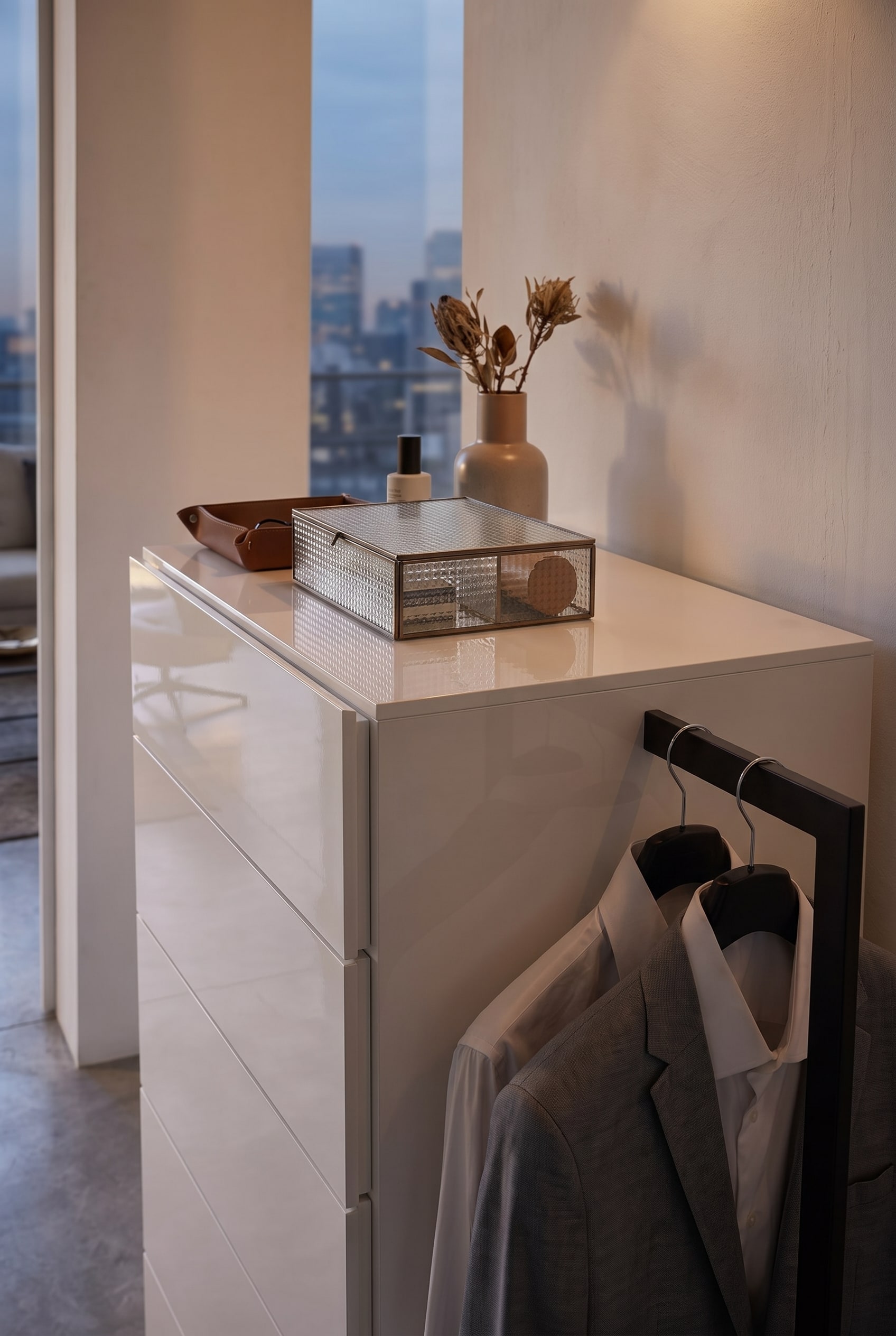 Detailaufnahme einer weißen Kommode mit geriffelter Glasbox, Lederschale und einer seitlichen Kleiderstange mit grauem Sakko.