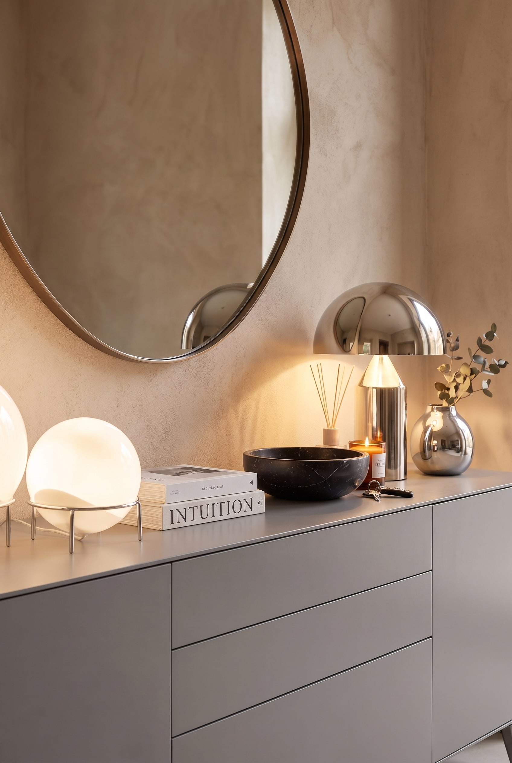 Detailaufnahme der Sideboard-Dekoration mit zwei runden, leuchtenden Tischlampen, einer schwarzen Marmorschale und B&uuml;chern auf matter Oberfl&auml;che.
