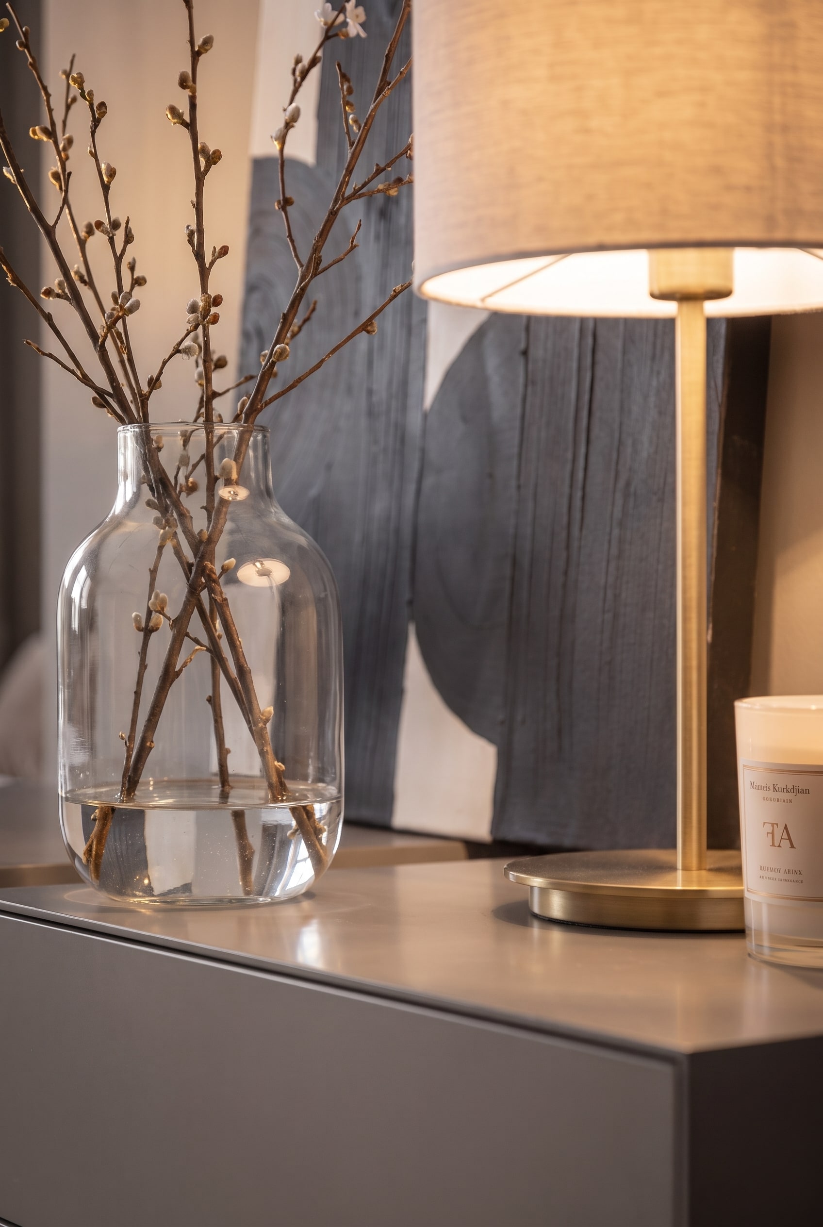 Detailansicht einer bauchigen Glasvase mit Zweigen neben einer leuchtenden Tischlampe und einer Kerze auf einer Kommode.