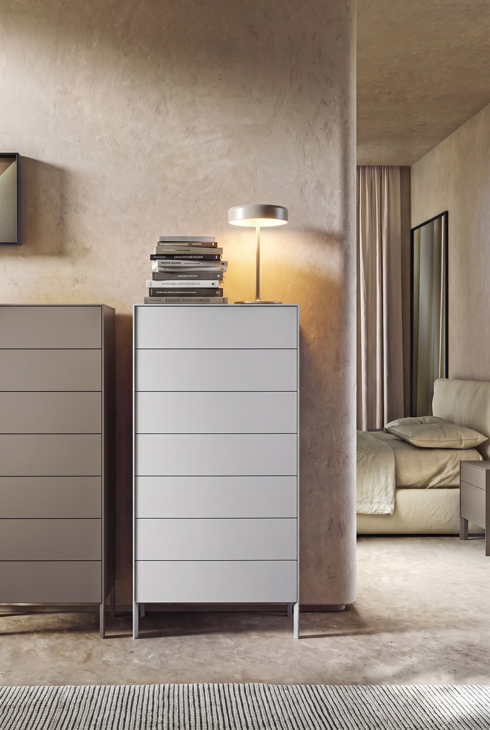 Minimalistisches Schlafzimmer mit hoher grauer Kommode, auf der linken Seite massiver B&uuml;cherstapel und Tischleuchte, rechte Seite leer