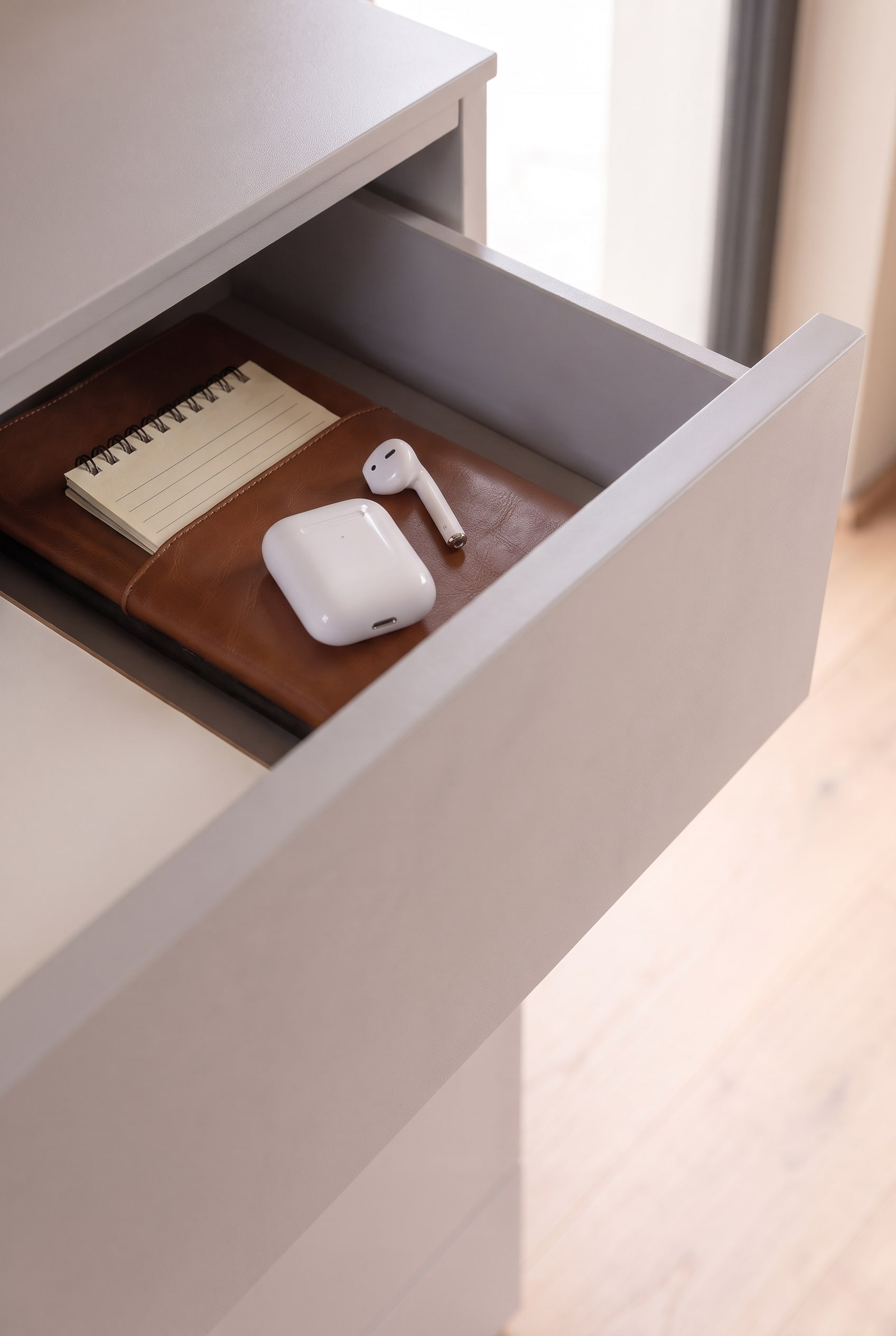Ge&ouml;ffnete Schublade einer greigen Kommode mit einem Leder-Notizbuch und wei&szlig;en AirPods.